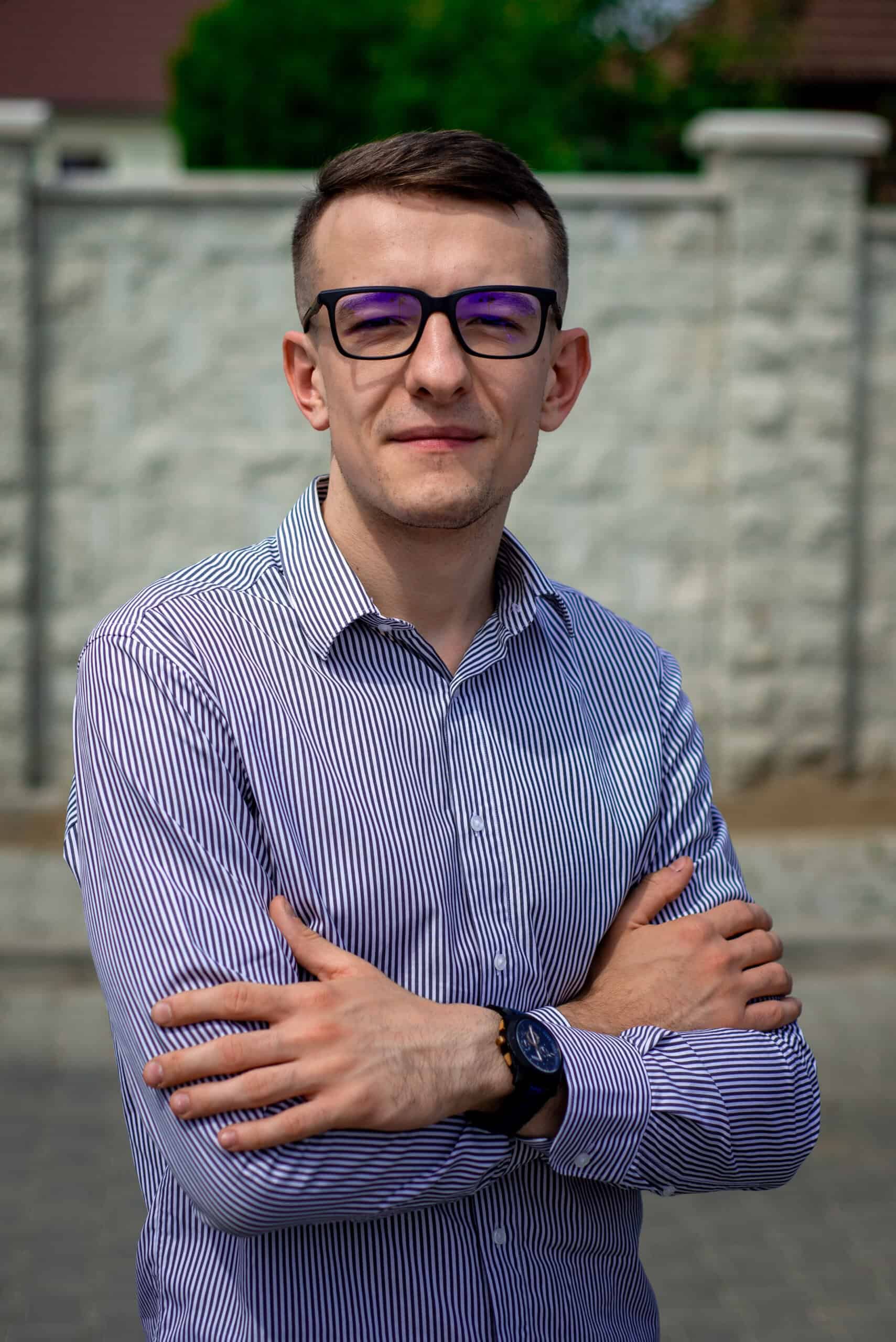 Tânăr profesionist în fața unui zid de beton, purtând o cămașă cu dungi și ochelari, exprimând încre.