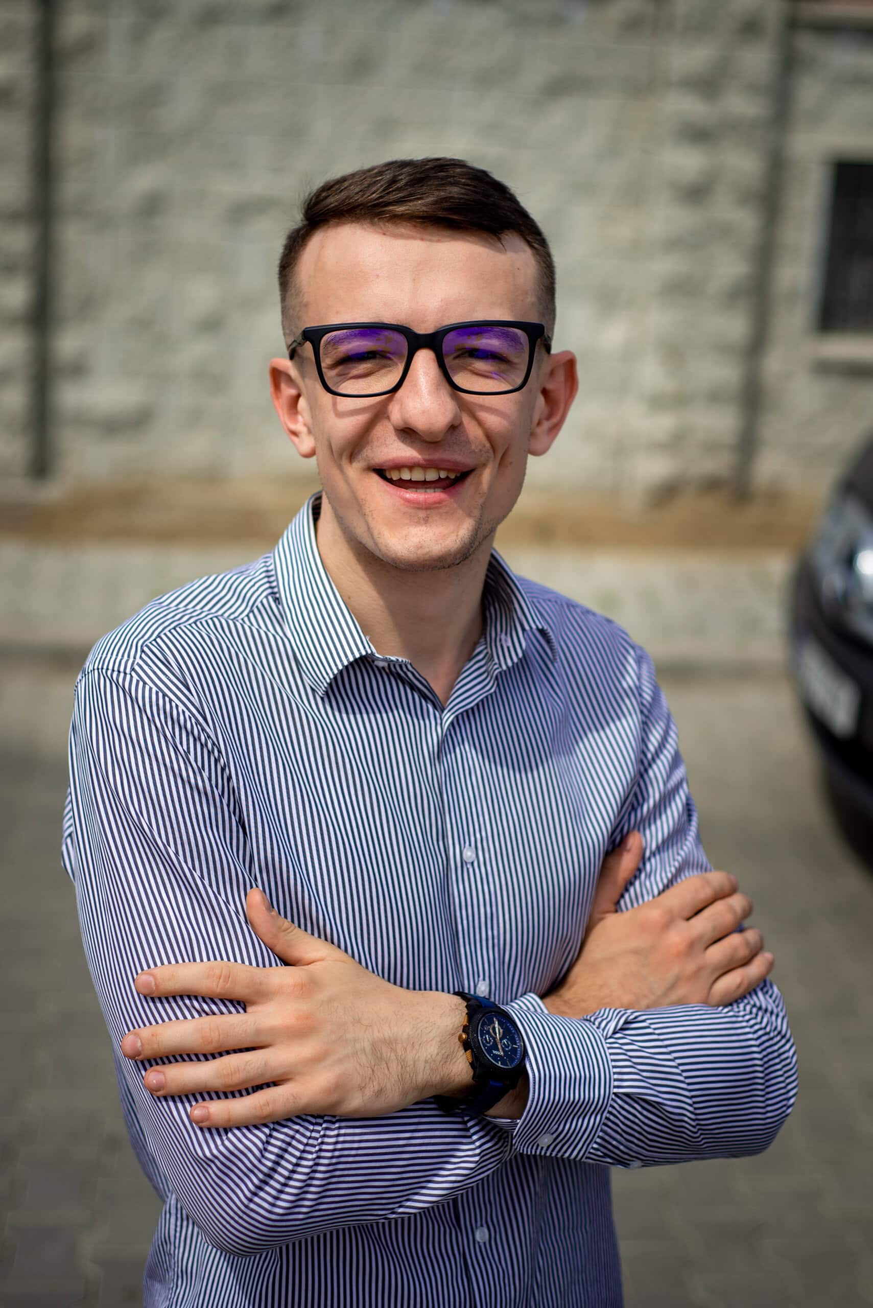 Tânăr bărbat cu ochelari, zâmbind în aer liber, purtând cămașă cu dungi și ceas, în fața unei clădir.